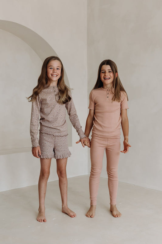 Two girls standing hand-in-hand against a soft beige backdrop, both smiling brightly. One wears a mauve floral-print long sleeve top with matching ruffled shorts, while the other wears a dusty pink ribbed short sleeve top and matching full-length leggings.