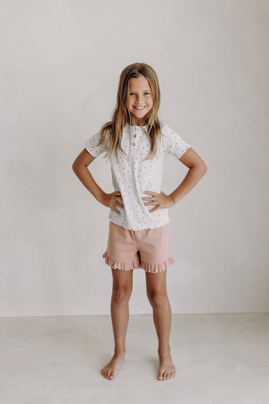 Young girl smiling confidently in a short-sleeved white ribbed top with a delicate floral print and blush pink ruffle shorts. She stands barefoot against a neutral background with hands on hips, showcasing a playful and comfy outfit.