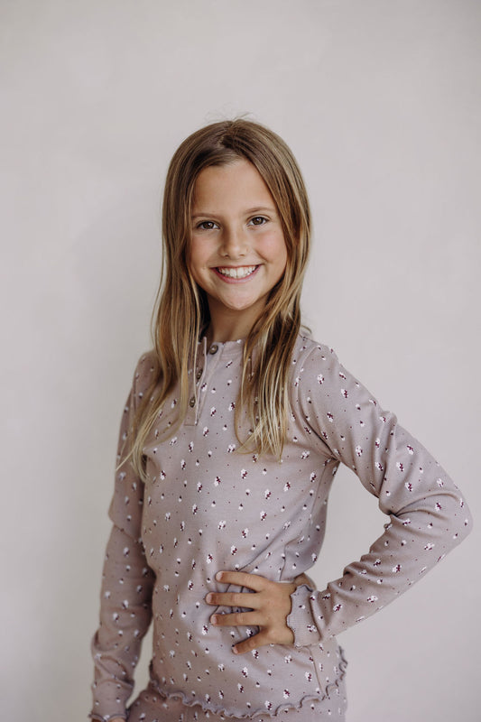 Smiling young girl stands confidently against a plain background, wearing a soft mauve long-sleeve henley top and matching shorts with a delicate floral print. One hand rests on her hip, highlighting the relaxed, playful vibe of the loungewear set.
