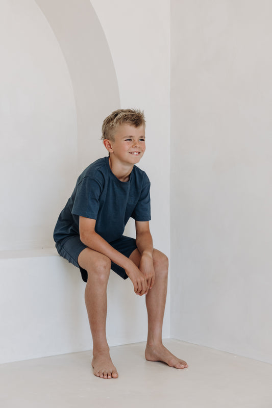 Young boy in a navy blue loungewear set, sitting playfully in a white alcove with a relaxed smile. The matching short-sleeved top and shorts are part of the Inspired by Nature kids' collection, designed for comfort and movement.