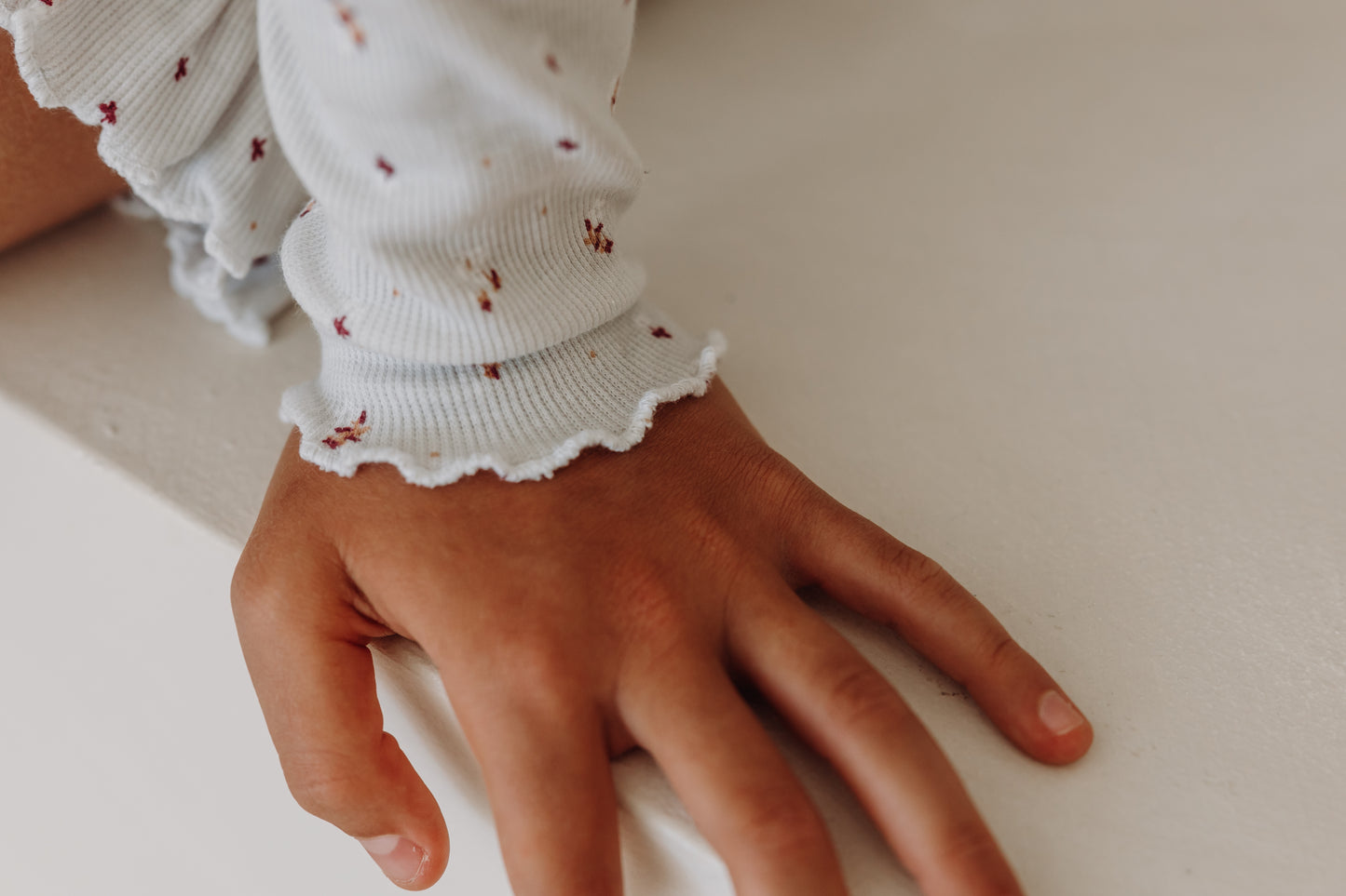 Close-up of a child's hand resting on a white surface, wearing a long-sleeve top with a white ribbed texture and a scattered burgundy floral print. The sleeve is finished with a soft ruffled edge.