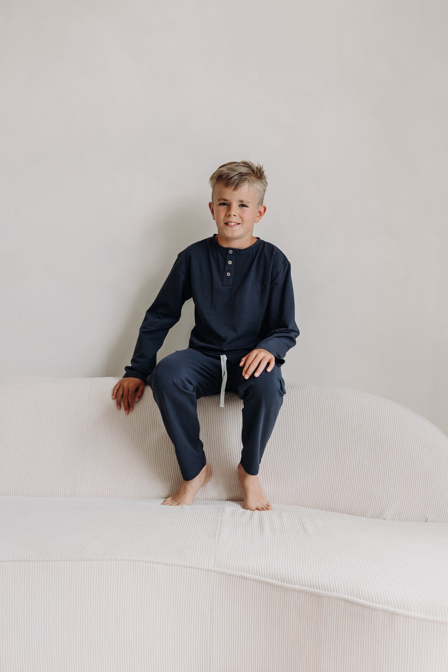 Young boy sitting on a cream couch wearing Inspired by Nature SA's navy long sleeve pyjama set made from organic cotton. Designed for softness, comfort, and eco-friendly sleepwear style.
