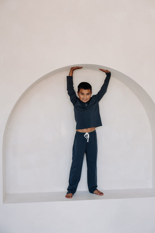 Smiling child wearing Inspired by Nature SA navy blue organic cotton pyjama set with long sleeves and drawstring pants. Standing barefoot inside a modern white arched alcove posing playfully in soft sustainable sleepwear.