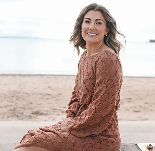 Woman sitting on a beach in a textured, rust-colored sweater dress, smiling at the camera with a calm ocean and soft sky in the background.